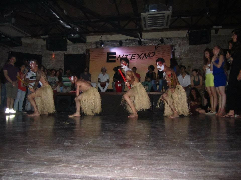 Four performers in grass skirts and white face paint dancing maculelê with sticks in front of an audience in Limassol
